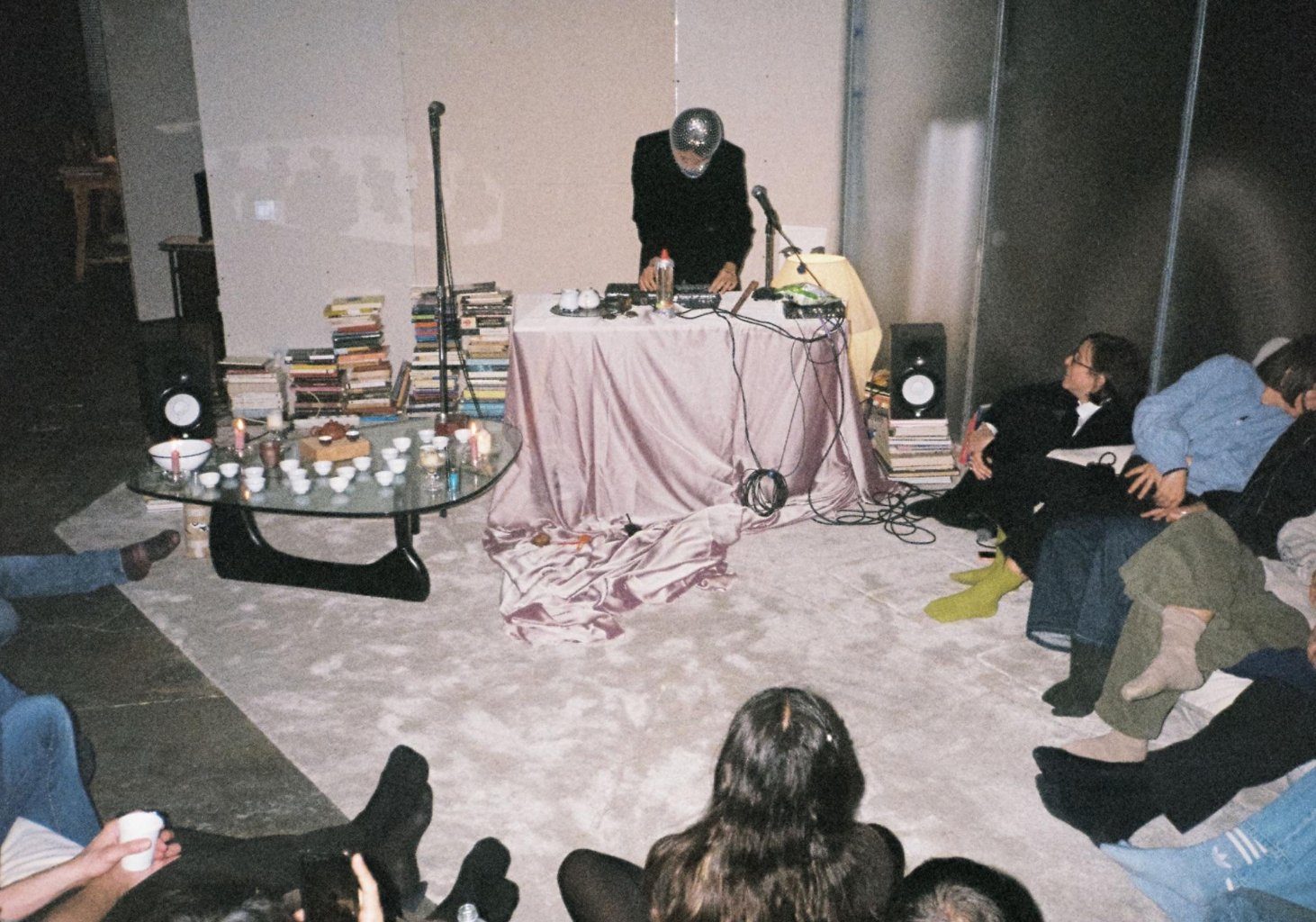 kuanmaru performing a tea ceremony electronic concert, with audience seated on the floor around a pink-draped table of teaware and electronics